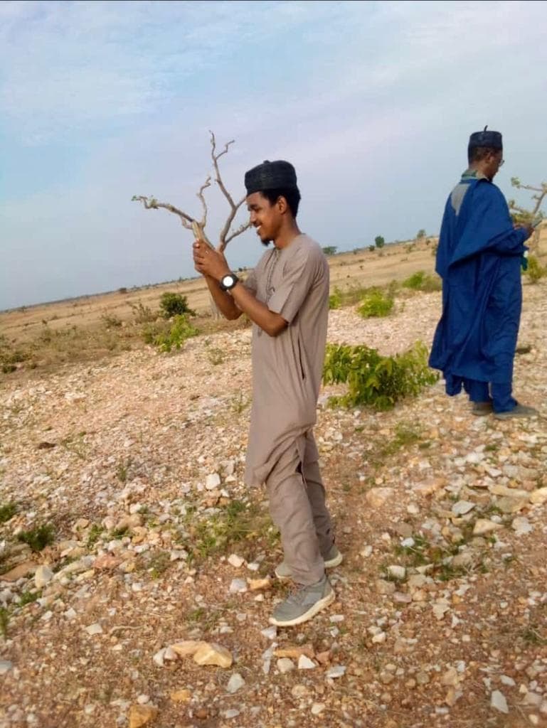 Maharaz holding a dry branch on stony savannah ground, with another man in blue in the distance